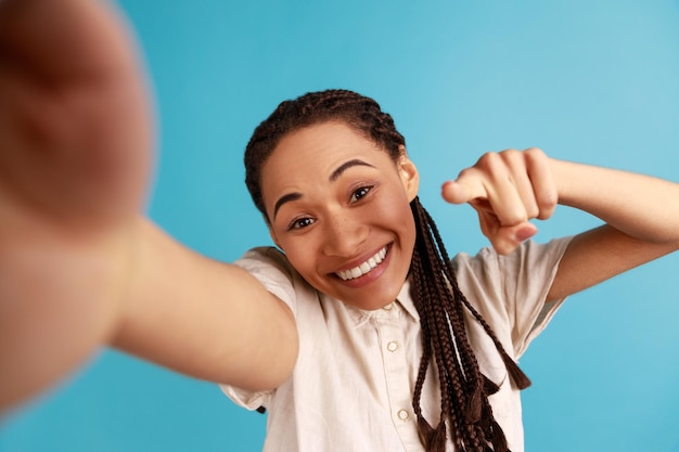 Vrolijke vrouw met zwarte dreadlocks heeft tevreden gezichtsuitdrukking maken selfie spreekt oprechte emoties wijzende vinger naar camera POV Indoor studio opname geïsoleerd op blauwe achtergrond