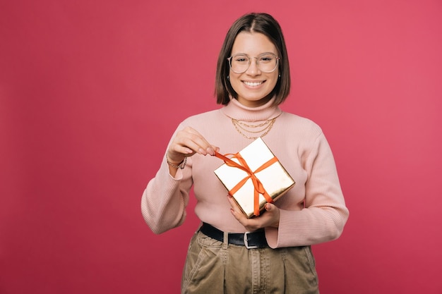 Vrolijke vrouw pakt een geschenkdoos uit terwijl ze naar de camera glimlacht Studio-opname over roze achtergrond