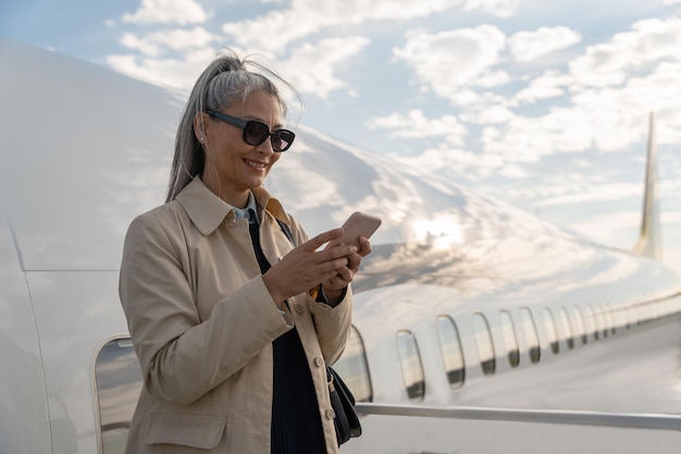 Vrolijke vrouw passagier met behulp van telefoon permanent buiten op de luchthaven in de buurt van vliegtuig