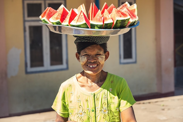 Vrolijke vrouw uit Birma (Myanmar) met een groot bord gesneden watermeloenen op haar hoofd.