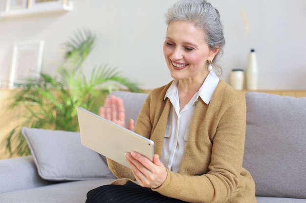 Vrolijke vrouw van middelbare leeftijd die op de bank zit, computertablet-apps gebruikt, naar het scherm kijkt, goed nieuws leest in een sociaal netwerk, online winkelt of chat.