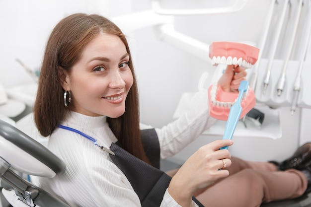 Vrolijke vrouwelijke patiënt glimlachen, tandheelkundige model en tandenborstel houden op tandartsstoel