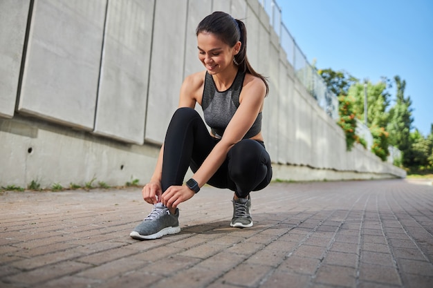 Vrolijke vrouwelijke persoon die op het trottoir staat terwijl ze schoenveters strikt tijdens de ochtendtraining