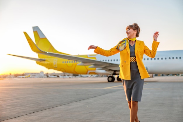 Vrolijke vrouwelijke stewardess in luchtvaartuniform die dansbewegingen doet en glimlacht terwijl ze buiten op het vliegveld staat