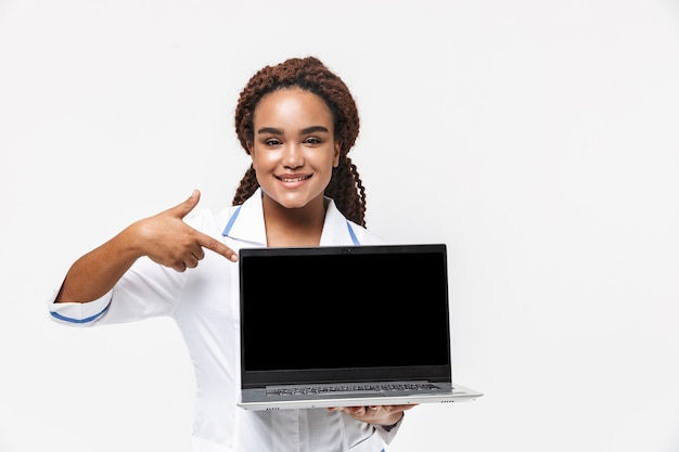 Vrolijke vrouwelijke verpleegster met een medische jas die een laptop vasthoudt die tegen een witte muur wordt geïsoleerd