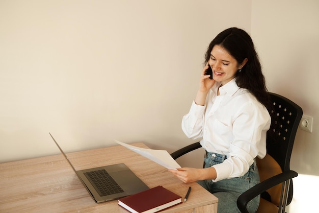 Vrolijke vrouwenmanager die telefonisch praat en aan een laptop werkt De baas van het meisje werkt op kantoor