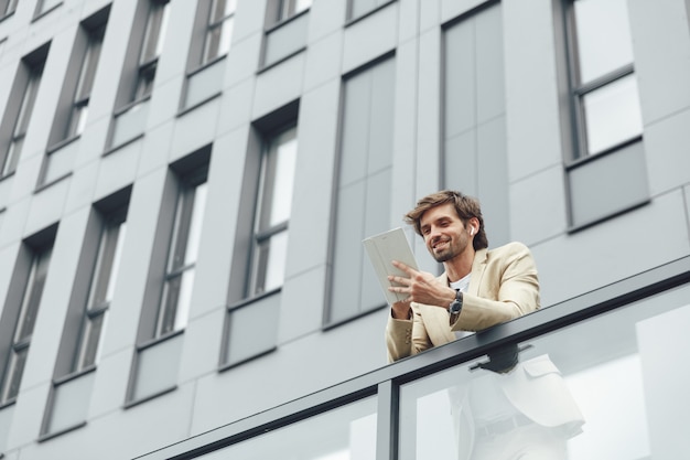 Vrolijke zakenman in formele kleding die online aan digitale tablet werkt terwijl hij buiten op het balkon van het kantoorgebouw staat