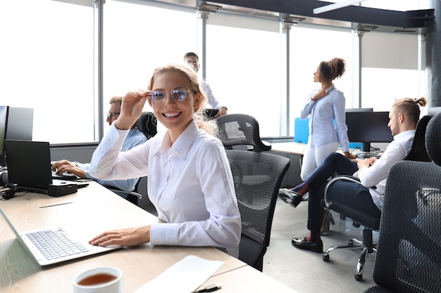 Vrolijke zakenvrouw in formalwear die op laptop in het moderne kantoor werkt met collega's op de achtergrond.