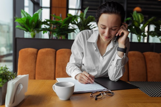 Vrolijke zakenvrouw praten op smartphone en het maken van aantekeningen in notitieblok aan tafel in café