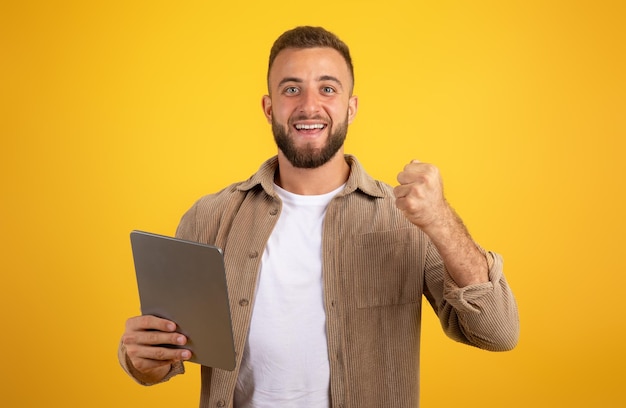 Vrolijke, zelfverzekerde knappe Europese duizendjarige man met baard gebruikt tablet en viert overwinning met vuist