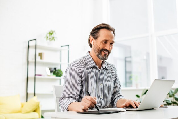 Vrolijke zelfverzekerde man van middelbare leeftijd zit aan de tafel en kijkt naar het scherm van zijn laptop terwijl hij een stylus in zijn rechterhand houdt