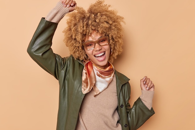 Foto vrolijke zorgeloze vrouw met krullend haar danst en viert iets houdt de armen omhoog, gekleed in modieuze kleding geïsoleerd op een beige achtergrond, voelt zich gelukkig en ontspannen. gelukkige emoties