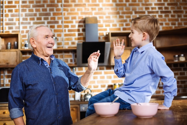 Vrolijke zorgzame oudere man met plezier met zijn kleinzoon terwijl hij in de keuken staat