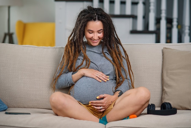 Vrolijke zwangere vrouw met dreadlocks zit op een comfortabele bank in de woonkamer en voelt zacht aan