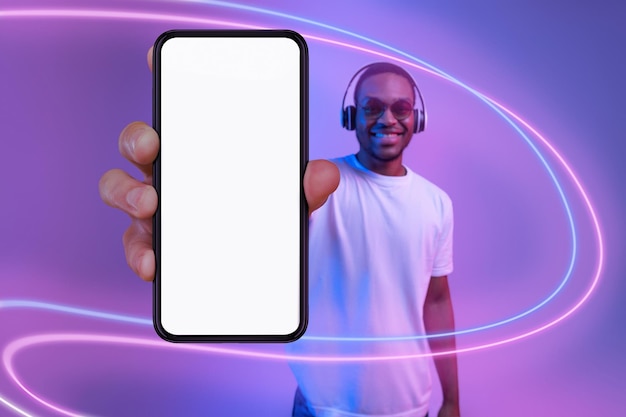 Vrolijke zwarte man die mobiele telefoon laat zien met behulp van een mockup voor draadloze hoofdtelefoons