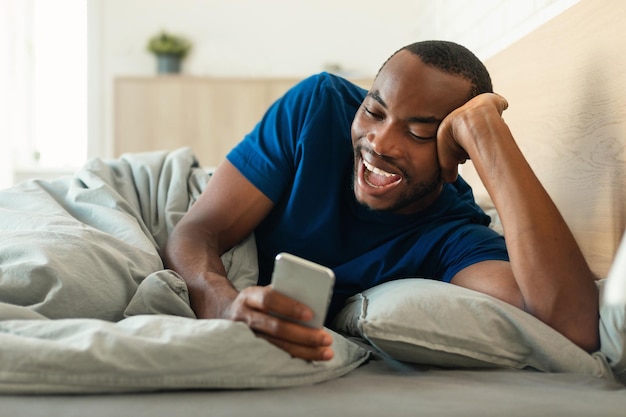 Vrolijke zwarte man met smartphone liggend in bed in slaapkamer