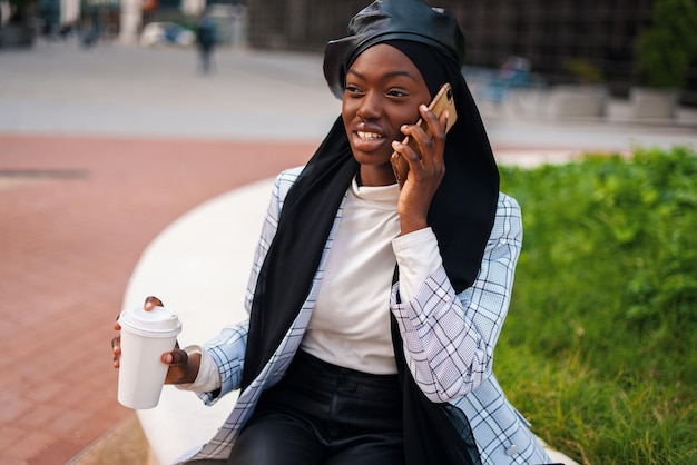 Vrolijke zwarte vrouw met afhaalkoffie die op smartphone spreekt