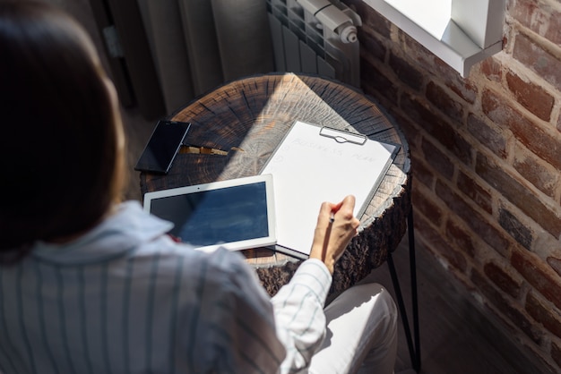 Vrouw aan de tafel voor het raam met een tablet en een notebook, businessplan, freelance, opstarten