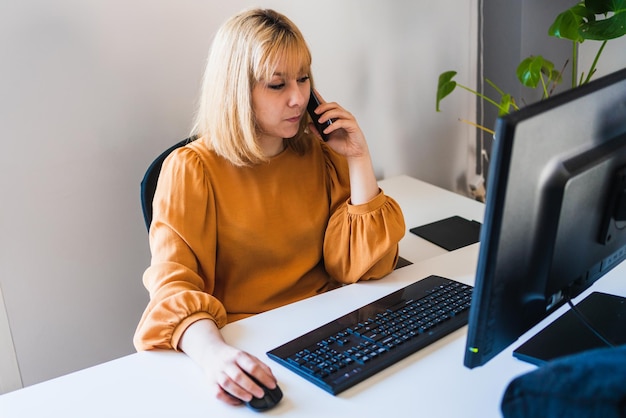 Vrouw aan het werk praten op haar smartphone voor een desktopcomputer op kantoor