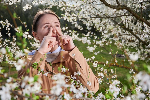 Vrouw allergisch lijdend aan seizoensgebonden allergie in de lente poseert in een bloeiende tuin in de lente Jonge vrouw niezen voor een bloeibare boom Voorjaarsallergie concept