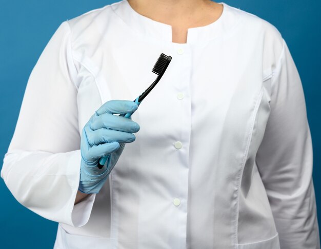 Vrouw arts in een witte medische jas en masker staat op een blauwe muur en houdt een plastic tandenborstel, close-up