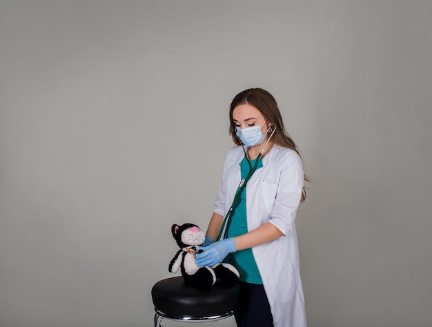 vrouw arts in handschoenen masker en witte jas met een stethoscoop luistert naar een stuk speelgoed op een grijze backgro