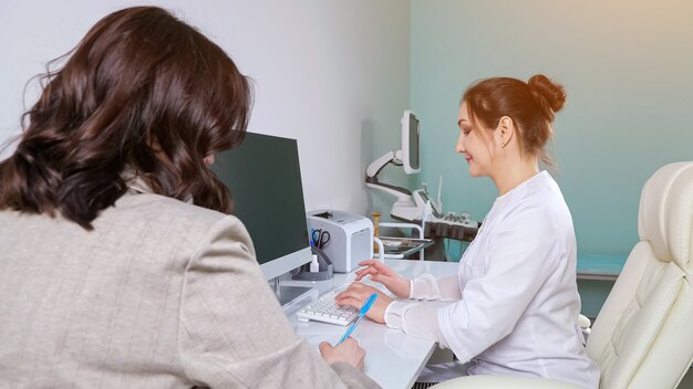 Vrouw arts in witte uniform typen informatie op computertoetsenbord luisteren naar vrouwelijke patiënt in ziekenhuis kantoor op afspraak close view