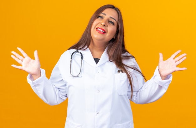 Vrouw arts van middelbare leeftijd in witte jas met stethoscoop, blij en positief lachend, opheffende armen die over oranje muur staan