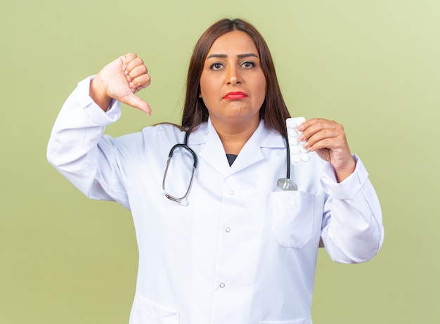 Vrouw arts van middelbare leeftijd in witte jas met stethoscoop met blaar met pillen die er ontevreden uitziet met duimen naar beneden staande over groene muur