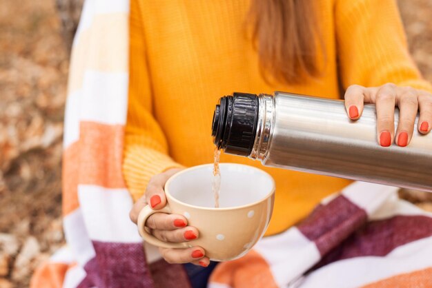 Vrouw bedekt met warme plaid die hete thee uit de thermosfles giet in de gele beker