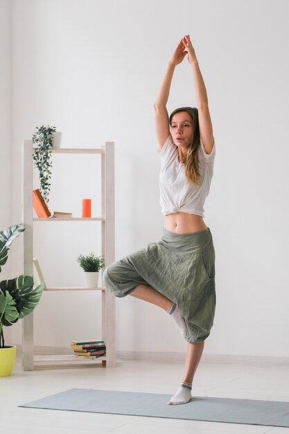 Vrouw beoefent yoga. Boom pose staande op mat binnenkant van gezellige kamer met planten en groen. Gezond levensstijlconcept. Ruimte kopiëren.