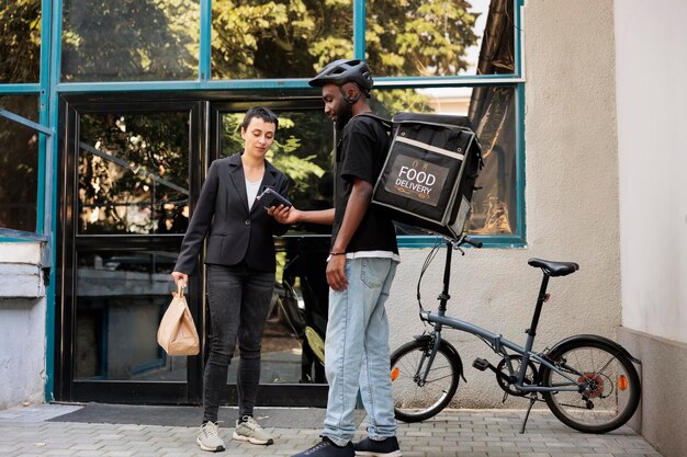 Vrouw betaalt voor bezorgservice voor kantoormaaltijden met creditcard, contactloze pos-terminalbetaling. Afro-amerikaanse koerier die afhaalmaaltijden aflevert aan bedrijfsmedewerker, breed schot