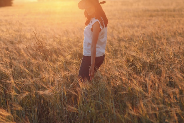 Vrouw boer in cowboyhoed wandelen met de handen op de oren op landbouwgebied op zonsondergang