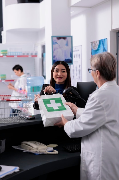 Vrouw chatten met apotheekkassier bij kassa, boodschappentas met medische producten meenemen. Oudere drogisterijmedewerker die jonge Aziatische klantmedicijnen online bestelpakket geeft aan de balie