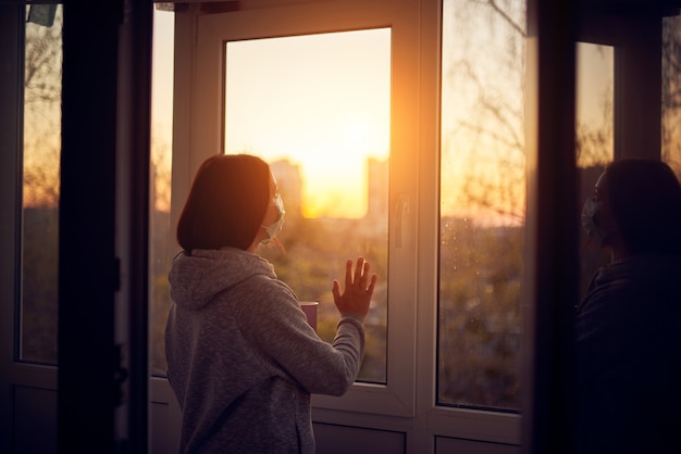 Vrouw dichtbij venster bij zonsondergang afzonderlijk voor virusuitbraak thuis. Blijf thuis concept