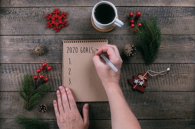 Vrouw die 2020-doelstellingen in notitieboekje met vakantiedecoratie schrijven op houten lijst
