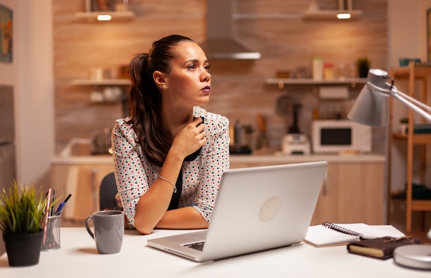 Vrouw die aan haar carrière denkt terwijl ze 's avonds laat in de keuken aan een deadline werkt. Werknemer die om middernacht moderne technologie gebruikt en overuren maakt voor werk, zaken, drukte, carrière, netwerk, levensstijl