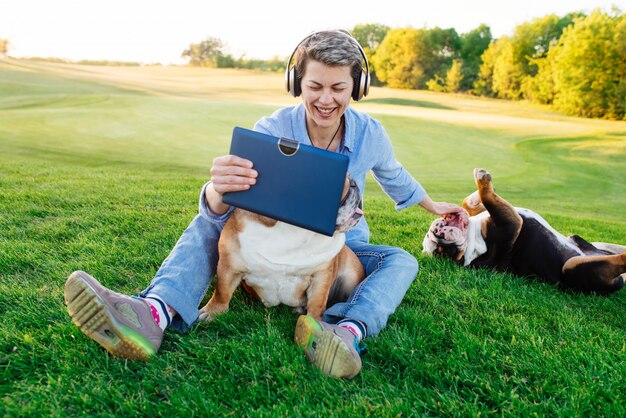 Vrouw die aan muziek of audioboek luistert, video op tablet bekijkt en op weide met twee honden / buldoggen in het park rust