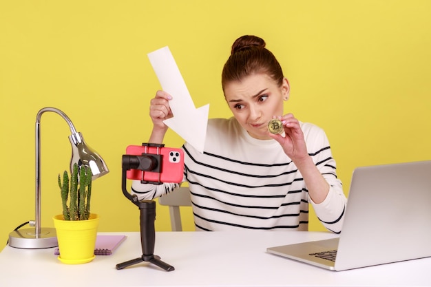 Vrouw die bitcoin en witte pijl toont die naar beneden wijst terwijl ze op de werkplek zit en video opneemt