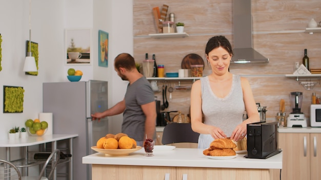 Vrouw die broodrooster gebruikt om brood in de keuken te roosteren tijdens het ontbijt. Jonge huisvrouw die thuis een ochtendmaaltijd kookt, vrolijk met genegenheid en liefde