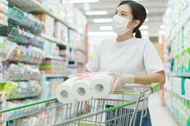 Vrouw die chirurgisch masker en handschoenen draagt, die toiletpapierbroodje in supermarkt koopt. Paniek winkelen na een pandemie van het coronavirus.