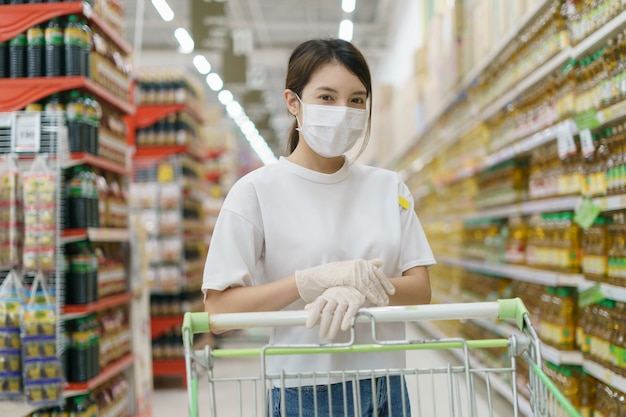 Vrouw die chirurgisch masker en handschoenen met een boodschappenwagentje draagt, dat tijdens een pandemie van coronavirus winkelt.