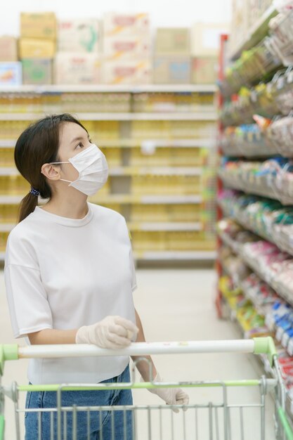 Vrouw die chirurgisch masker en handschoenen met een boodschappenwagentje draagt, dat tijdens een pandemie van coronavirus winkelt.