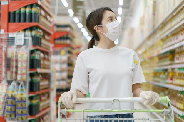 Vrouw die chirurgisch masker en handschoenen met een boodschappenwagentje draagt, dat tijdens een pandemie van coronavirus winkelt.