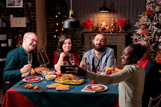 Vrouw die de groepsfoto van de kerstviering van vrienden op mobiele telefoon neemt, divers mensenportret aan feestelijke eettafel. Grote familiebijeenkomst, kerst vieren, traditioneel eten, drinken