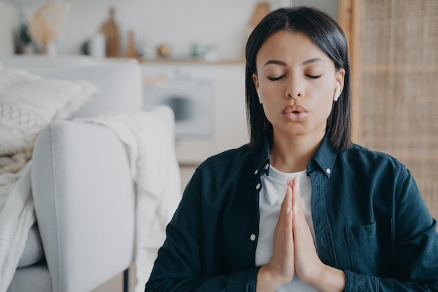 Vrouw die diep ademhaalt, luistert naar meditatie in oortelefoon met namaste-gebaar thuis Stressverlichting