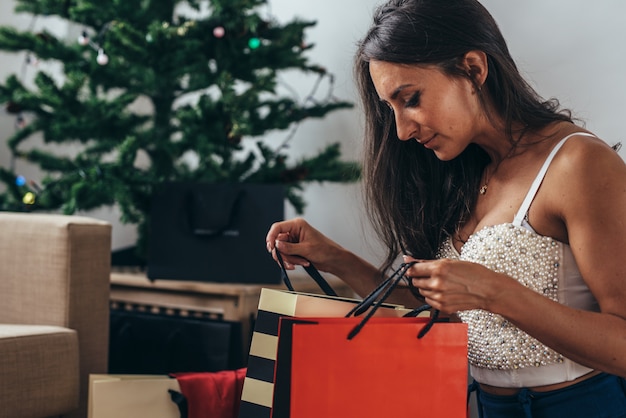 Vrouw die door de Kerstmisboom situeert en een heden neemt uit het winkelen zak