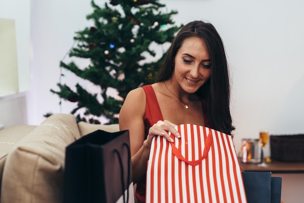 Vrouw die door de Kerstmisboom situeert en een heden neemt uit het winkelen zak