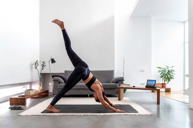 Vrouw die één been naar beneden toegekeerde hond doen splitsen thuis gebruikend online yogatrainingprogramma in de tablet