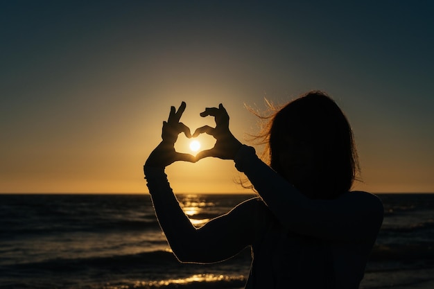 Vrouw die een hart maakt met haar vingers tegen het licht terwijl de zon ondergaat